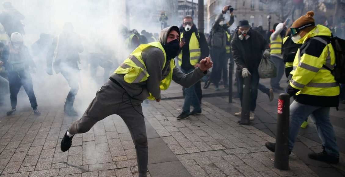A protestor wearing a yellow vest (gilet jaune) throws a tear gas canister back at police during a protest in Paris