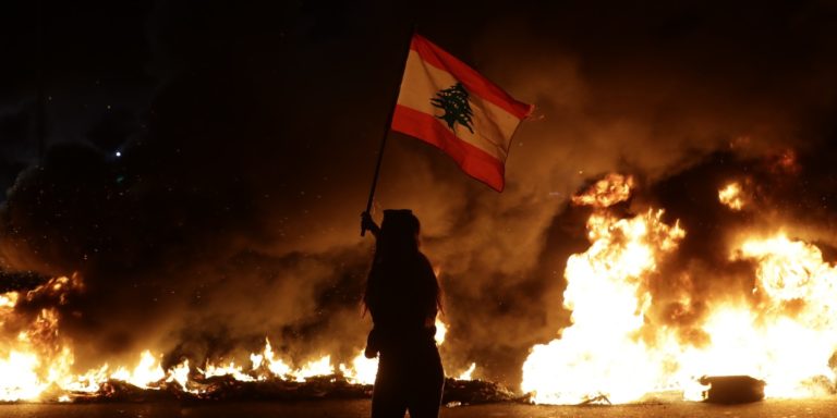 An anti-government protester waves a Lebanese flag in front of burning tires that block the main highway linking the cities of Tripoli and Beirut on Nov. 13, 2019