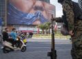 An armed Iranian police officer holding a rifle monitors the area as motorcyclists ride beneath a billboard depicting an AI-generated image of the Strait of Hormuz and an effigy of US President Donald Trump, displayed on the wall of a state building in downtown Tehran, Iran, on May 3, 2026.