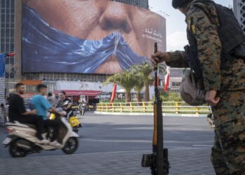 An armed Iranian police officer holding a rifle monitors the area as motorcyclists ride beneath a billboard depicting an AI-generated image of the Strait of Hormuz and an effigy of US President Donald Trump, displayed on the wall of a state building in downtown Tehran, Iran, on May 3, 2026.