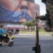 An armed Iranian police officer holding a rifle monitors the area as motorcyclists ride beneath a billboard depicting an AI-generated image of the Strait of Hormuz and an effigy of US President Donald Trump, displayed on the wall of a state building in downtown Tehran, Iran, on May 3, 2026.